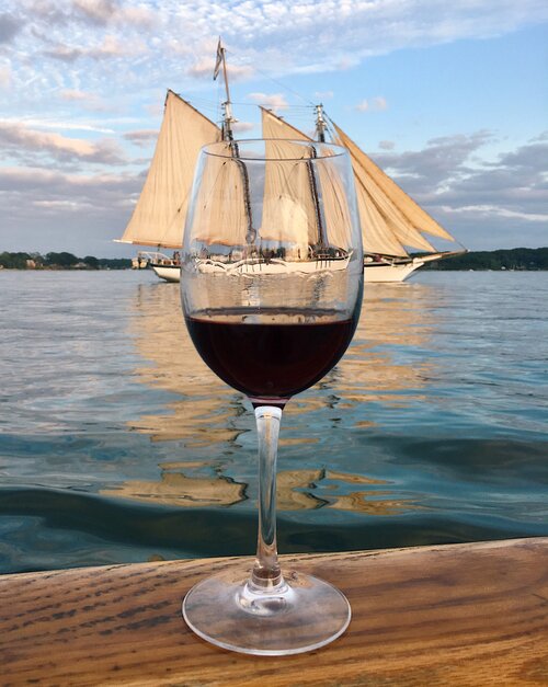 A double masted schooner passes behind a glass of red wine sitting on a dock in Portland, Maine